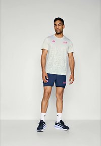 Hellgraues Sport-T-Shirt mit rosa Akzenten und gemustertem Design, kombiniert mit einer marineblauen Shorts. Weiße Socken und schwarze Sportschuhe mit blauen Akzenten.