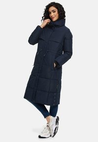 Marinblå pufferjacka med hög krage, elastiska dragsnören i midjan, quiltat mönster och sidofickor. Kombineras med denim och kraftiga sneakers.