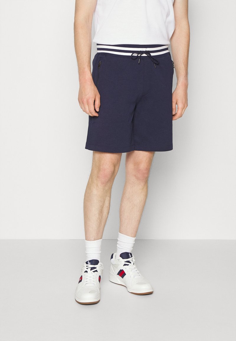 Navy blue shorts with white and navy striped waistband, drawstring, and side zip pockets, worn with white sneakers and crew socks.