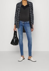 Camisa de cuadros en blanco y negro, camisola de encaje negra, vaqueros ajustados azules, zuecos blancos y bolso negro con textura. Diseño de conjunto sencillo y moderno.