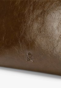 Producto de cuero marrón con una superficie texturizada, que presenta un detalle en relieve de calavera y tibias cruzadas. El material parece suave y flexible.