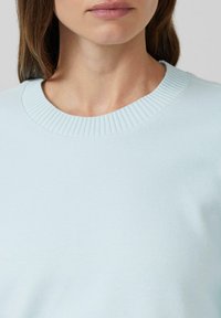 Kobieta ubrana w jasnoniebieski sweter o prą�żkowanym wzorze z okrągłym dekoltem, pokazujący szyję i górną część klatki piersiowej.