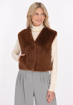 Gilet in finta pelliccia marrone con scollatura a V, caratterizzato da una morbida texture e due tasche laterali, indossato sopra un dolcevita beige e pantaloni grigi a pieghe.