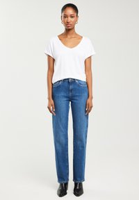 Kobieta ubrana w białą koszulkę z dekoltem w serek, niebieskie jeansy o prostych nogawkach, czarne botki za kostkę i złote kolczyki koła, stojąca na jednolitym tle.