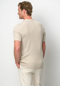 Beige kortärmad polo skjorta med en texturerad ribbad mönster och mjukt tyg. Slät fåll och krage, bärs med ljusa byxor.