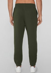 Dunkelgrüne Sweatpants mit einer schmal zulaufenden Beinform, elastischem Bund und seitlichen Taschen, kombiniert mit weißen Sneakers. Glatte Textur.