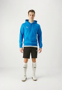 Sweatshirt azul com capuz e bolso canguru, calções pretos, meias brancas até ao joelho e ténis brancos com solas castanhas. Detalhes do logótipo em branco.