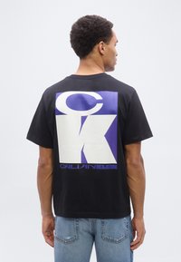 Mand iført sort Calvin Klein T-shirt med stort hvidt og lilla CK-logo på ryggen, kombineret med blå jeans, vendt væk.