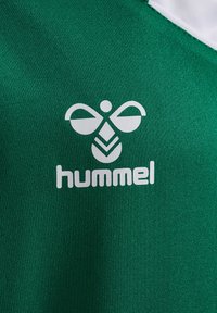 Jersey deportivo de tela verde con un logo blanco de diseño simétrico encima del nombre de la marca "hummel" en letras negritas.