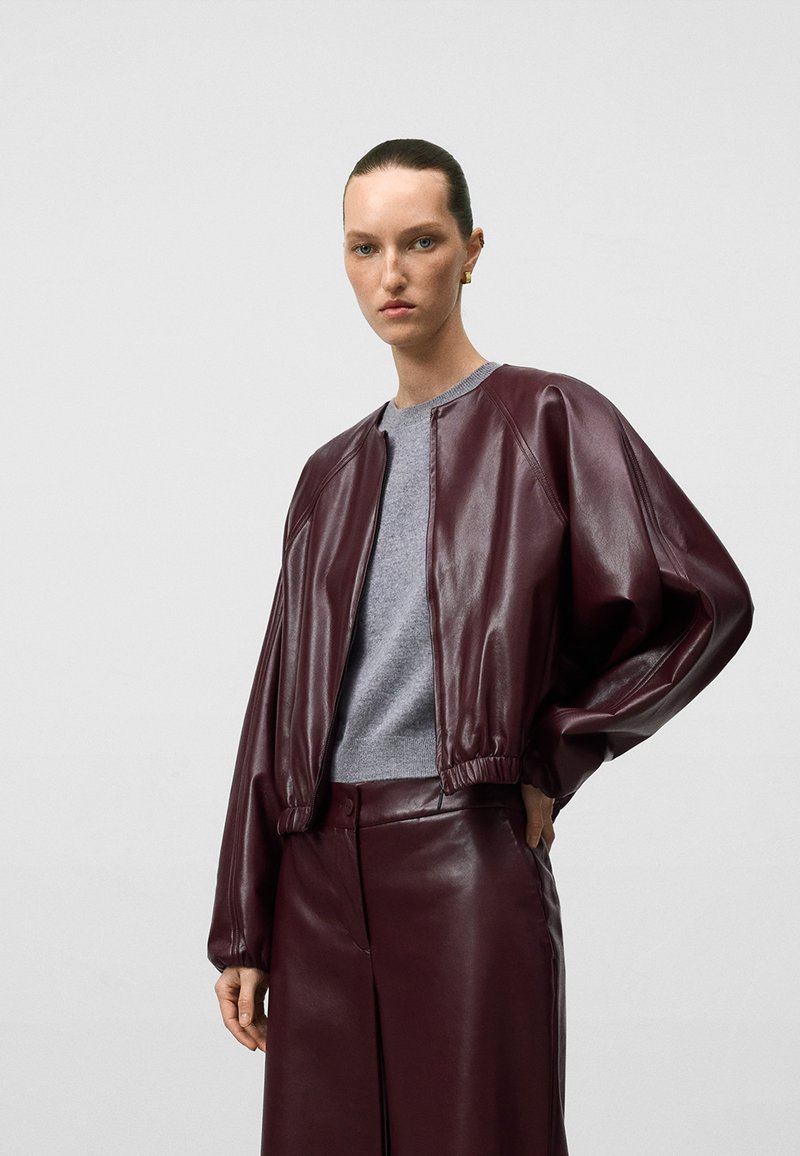 Giubbotto bomber in pelle bordeaux con polsini ed orlo elasticizzati, abbinato a un maglione grigio. Texture liscia e vestibilità rilassata in tutto il capo.