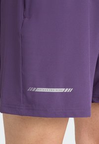Lila Sportshorts aus leichtem Stoff, mit einem reflektierenden "Adidas Training"-Logo in der Nähe des Saums und einer glatten Textur.