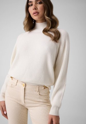 Vrouw met golvend haar die een witte geribde coltrui en lichtbeige broek met patroon draagt tegen een effen grijze achtergrond.
