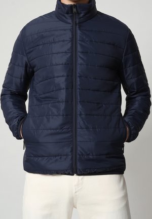 Mann trägt eine marineblaue gesteppte Jacke mit Frontreißverschluss und Händen in den Seitentaschen, kombiniert mit weißen Hosen.