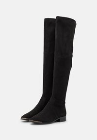 Botas negras por encima de la rodilla hechas de ante, con punta puntiaguda, tacón bajo y detalle metálico en la puntera. Textura lisa y diseño ajustado.