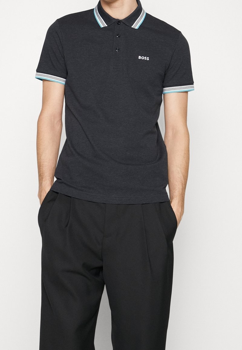 Homme portant un polo gris foncé avec un col et des bords de manches rayés blanc, bleu et gris, avec le logo "BOSS" sur la poitrine, et un pantalon noir.