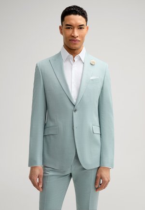 Costume bleu clair en tissu lisse avec une coupe ajustée. Présente un revers en pointe, une fermeture à un bouton et une épingle de revers florale.