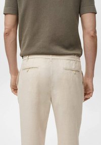 Beige linnen broek met een ontspannen pasvorm, voorzien van twee achterzakken met knopen en een elastische tailleband, gecombineerd met een gestructureerd olijfgroen shirt.