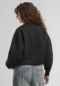 Felpa nera con scollo rotondo e spalle scese, abbinata a jeans azzurri chiari con un design ricamato decorativo sulla tasca.