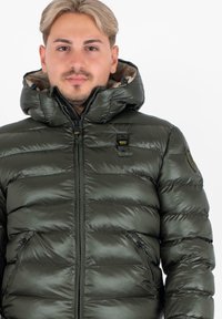 Jonge man in een glanzende donkergroene pufferjack met capuchon, rits dicht, poserend tegen een plain witte achtergrond.