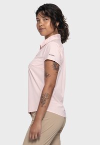 Light pink polo shirt with a collar, short sleeves, and a logo on the left sleeve. Smooth fabric, relaxed fit, and a side pocket detail.