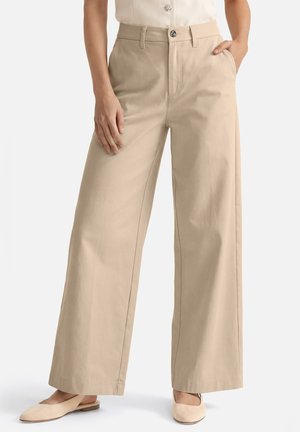 Femme portant un pantalon large beige et des chaussures plates assorties, une main dans la poche et l'autre reposant sur la cuisse.