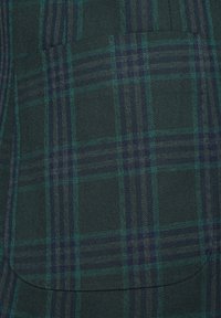 Zielona i granatowa tkanina w kratę o fakturowanym splocie. Posiada kieszeń oraz wyraźne linie wzoru przecinające się, tworząc warstwowy efekt.