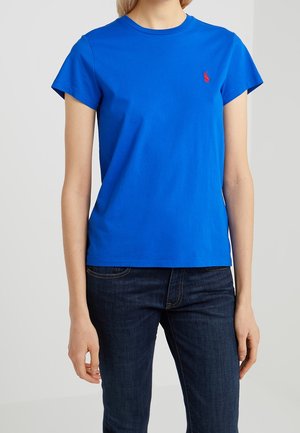 Person iført en lys blå T-shirt med korte ærmer og en lille rød logo på brystet samt mørkeblå jeans mod en ensfarvet baggrund.