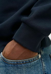 Marineblauwe sweater manchet met ribbeltextuur, gecombineerd met licht denim jeans, waarbij een hand die in de zak rust gedeeltelijk zichtbaar is.