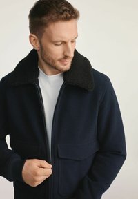 Granatowa kurtka wykonana z mieszanki wełny z czarnym kołnierzem z owczej skóry, zapinana na zamek, z dwiema przednimi kieszeniami. Gładka faktura i dopasowany krój.