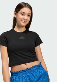 Crni kratkodlaki crop top s glatkom teksturom i suptilnim sivim Adidas logotipom na prsima, uparen s plavim kratkim hlačama sa leopard printom.