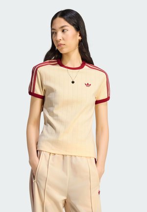 Ung kvinde iført cremefarvet Adidas t-shirt og bukser med røde striber, står med hænderne i lommerne og ser til siden.