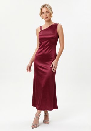 Femme portant une robe de soirée en satin bordeaux sans manches avec un décolleté asymétrique et des talons hauts argentés à brides, sur fond blanc.