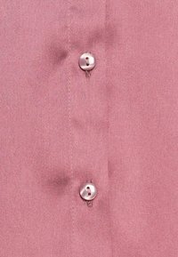 Camisa rosa de botão com textura de tecido suave, apresentando dois botões metálicos redondos e costura fina ao longo da pala.