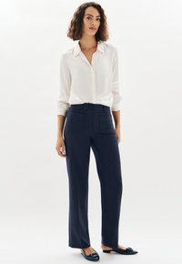 Femme debout portant une chemise blanche à boutons rentrée dans un pantalon droit bleu marine avec des poches plaquées à l'avant et des ballerines pointues bleues marines.