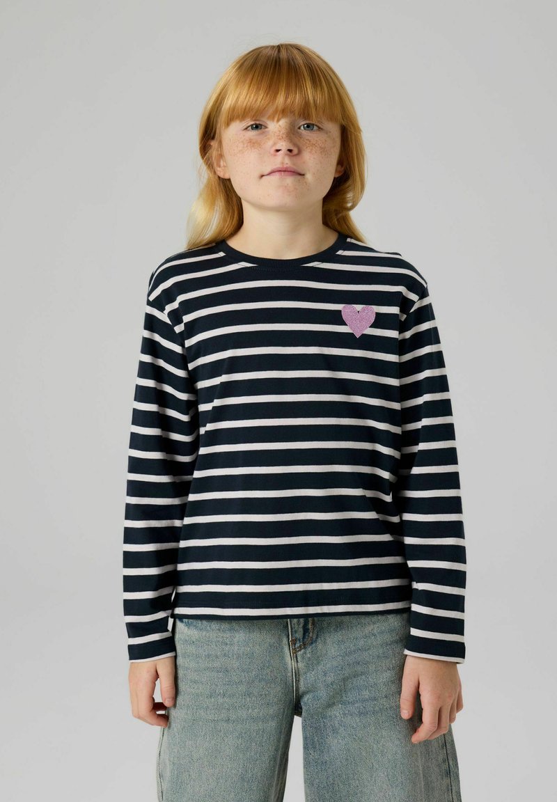 Schwarz-weiß gestreiftes Langarmshirt mit pinkfarbenem Glitzerherz auf der linken Brust, kombiniert mit hellblauen Jeans.