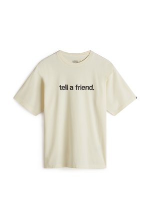 Henkilö, joka pukeutunut löysään kermaan valuvaan t-paitaan, jossa musta teksti "tell a friend." seisoo yhdellä kädellä tummien housujen taskussa.