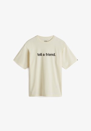 Personne portant un t-shirt ample crème avec le texte noir « tell a friend. » debout avec une main dans la poche d’un pantalon sombre.