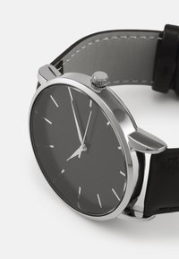 Črn ročni ura z elegantnim, okroglim ohišjem iz nerjavečega jekla, minimalistično črnim številčnico, srebrnimi kazalci in črnim usnjenim pasom.