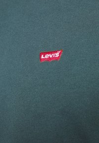 Grøn stof nærbillede med et lille rødt og hvidt Levi's-logo patch syet nær midten.