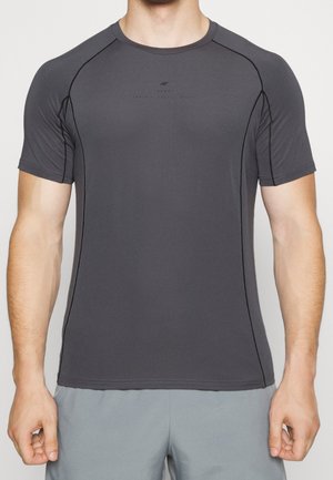 Atletyczny mężczyzna ubrany w dopasowaną ciemnoszarą koszulkę z krótkim rękawem z czarnymi przeszyciami oraz jasnoszare spodenki, stojący z luźno opuszczonymi ramionami.