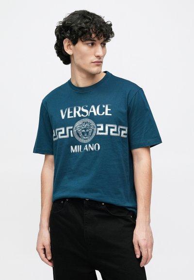 Purpura kokvilnas T-krekls ar sudraba grafisko logo, kurā attēlota Medūzas emblēma un grieķu raksts, saskaņots ar melnām džinsa biksēm.