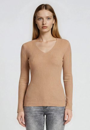 Donna bionda che indossa un maglione a coste beige a maniche lunghe con scollo a V e jeans grigi slavati, in piedi davanti a uno sfondo chiaro e uniforme.
