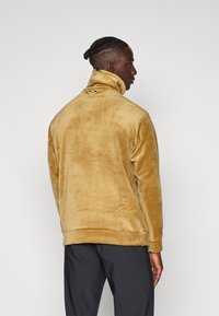 Sudadera de forro polar en un color amarillo apagado, con un cuello alto, tejido texturizado y una pequeña etiqueta con el logo en la parte posterior. Corte ajustado.