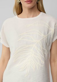 T-shirt en coton de couleur ivoire clair, arborant un grand motif de feuille texturée en crème brodé sur le devant, avec des manches courtes et un col rond.