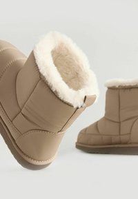 Stivaletti beige con un esterno texture, fodera in soffice pelliccia bianca, forma arrotondata e suola in gomma piatta. Presentano dettagli cuciti e una loop sul retro.