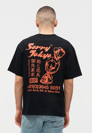 Lühikeste juustega mees, kes kannab musta T-särki, millel on suur oranž kommigraafika ja Jaapani tekst, seisab ühevärvilise heleda tausta ees.