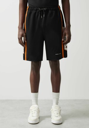 Man draagt zwarte sportshorts met oranje zijstrepen, witte sneakers en witte sportsokken, staand op een grijze vloer.