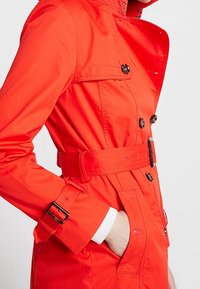 Jaskrawoczerwona trench coat z paskiem w talii, podwójnymi guzikami oraz zatrzaskami przy kołnierzu i mankietach. Gładki, lekki materiał.