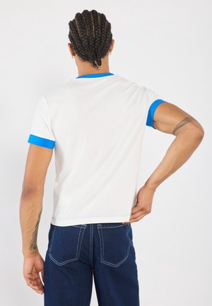 Osoba z warkoczami, ubrana w białą koszulę z niebieską lamówką i ciemne dżinsy, stojąca bokiem z jedną ręką na biodrze.