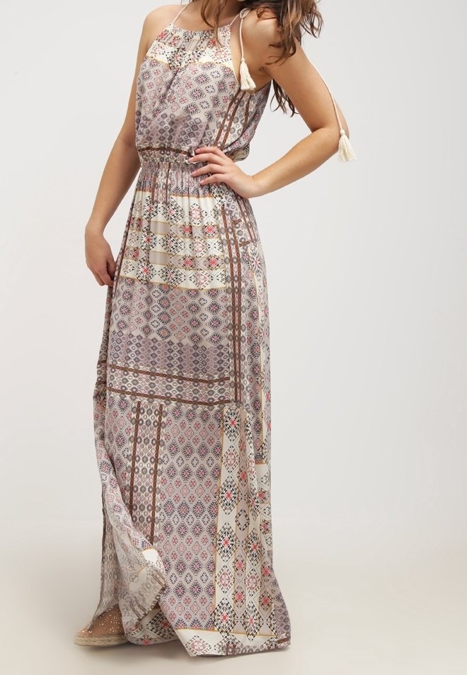 Femme portant une robe maxi sans manches à motifs avec des bretelles à pompons, debout avec une main sur la taille et l'autre détendue.